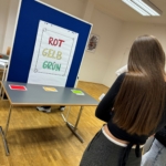 Eine Person mit langen braunen Haaren steht vor einem Tisch mit drei Karten - rot, gelb und grün. Hinter dem Tisch steht ein Schild auf einer blauen Tafel mit den deutschen Worten "ROT GELB GRÜN" in passenden Farben.