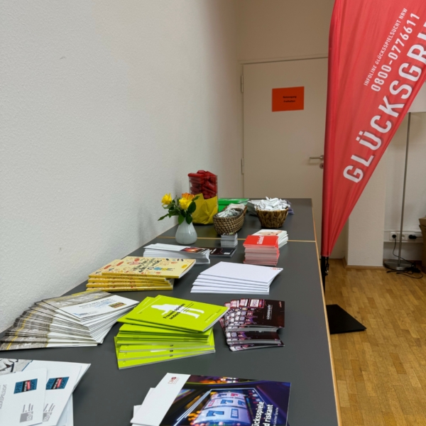 Ein Tisch mit bunten Broschüren, Heften, Snacks und einer kleinen Blumenvase steht vor einer weißen Wand. Eine rote Fahne mit der Aufschrift GLÜCKSOR steht im Hintergrund. Der Raum hat einen Holzfußboden.