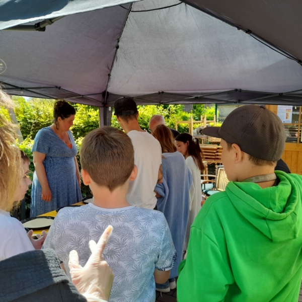 Eine Gruppe von Menschen, meist Kinder und einige Erwachsene, steht unter einem Vordach bei einer Veranstaltung im Freien. Eine Frau in einem blauen Kleid steht hinter einem Tisch, während andere ihr gegenüber stehen, sich unterhalten und in der Schlange warten. Im Hintergrund scheint das Sonnenlicht.
