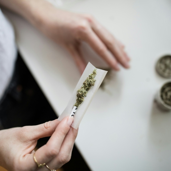 Eine Person hält ein mit gemahlenem Cannabis gefülltes Papier in der Hand und bereitet sich darauf vor, einen Joint zu drehen. Auf dem weißen Tisch im Hintergrund sind zwei Cannabis-Mahlwerke zu sehen.