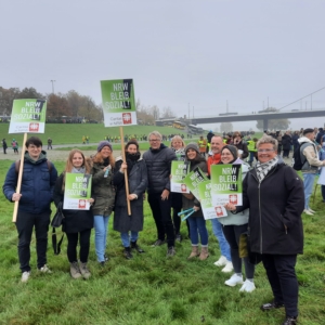 Eine Gruppe von Menschen steht auf einer Wiese zusammen und hält Schilder mit der Aufschrift "NRW BLEIB SOZIAL!" während einer öffentlichen Veranstaltung oder eines Protests im Freien. Im Hintergrund sind bei bewölktem Himmel weitere Menschen versammelt.