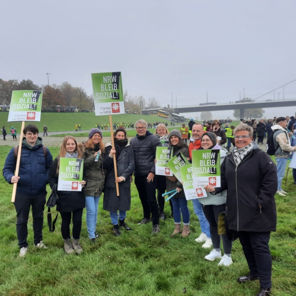Eine Gruppe von Menschen steht auf einer Wiese zusammen und hält Schilder mit der Aufschrift "NRW BLEIB SOZIAL!" während einer öffentlichen Veranstaltung oder eines Protests im Freien. Im Hintergrund sind bei bewölktem Himmel weitere Menschen versammelt.