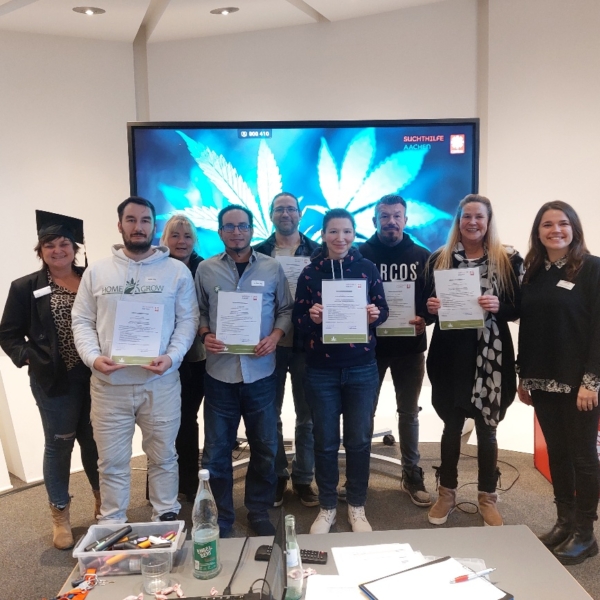 Eine Gruppe von acht Personen steht in einem Raum, lächelt und hält Urkunden in der Hand, während hinter ihnen ein großer Bildschirm ein Bild von Cannabisblättern zeigt. Im Vordergrund ist ein Tisch mit Papieren und Flaschen zu sehen.