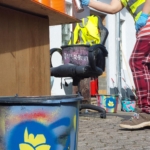 Eine Person in karierten Hosen und Handschuhen besprüht im Freien einen blauen Eimer mit einer gelben Blumenschablone. Orangefarbene Tabletts und eine Warnweste stehen auf einem Holztisch in der Nähe. Im Hintergrund sind weitere bemalte Eimer und ein Stuhl zu sehen.