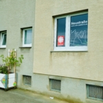 Ein beiges Gebäude mit drei kleinen Fenstern und einem größeren Fenster mit Schildern, darunter ein rotes Caritas-Logo. Blumenkästen und bunte Pflanzkübel mit Pflanzen schmücken die Außenwände und das Abflussrohr.