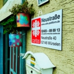 Ein Hauseingang mit einem bunten Blumentopf, einem weißen Briefkasten und einem Schild mit der Aufschrift Caritas Neustraße mit Kontaktinformationen und Adresse: Neustraße 40, 52146 Würselen. Über dem Schild steht die Zahl 40.