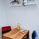 Ein kleiner Holztisch mit einem roten und einem blauen Stuhl, auf dem eine Glasflasche, ein Orangendrink, ein Glas und eine Zeitschrift stehen. Darüber steht ein weißes Regal mit Topfpflanzen vor einer schlichten weißen Wand.