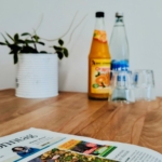 Ein Holztisch mit einer Zeitung, einer Topfpflanze, einer Glasflasche mit Wasser, einer Glasflasche mit Orangensaft und zwei leeren Gläsern steht an einer weißen Wand.