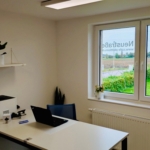 Ein helles, aufgeräumtes Büro mit einem weißen Schreibtisch, Laptop, Drucker und Büromaterial. Eine Pflanze steht auf einem Wandregal. Ein großes Fenster zeigt Grünflächen und Felder im Freien; auf dem Glas ist eine Beschilderung zu sehen.
