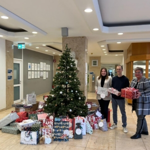Drei Personen stehen vor einem geschmückten Weihnachtsbaum in einer Bürohalle und halten eingepackte Geschenke in der Hand. Zahlreiche Geschenke sind um den Baum herum angeordnet und schaffen eine festliche Weihnachtsatmosphäre.