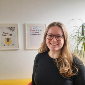 Eine Frau mit langen blonden Haaren und Brille steht lächelnd in einem Wohnzimmer mit gerahmter Kunst an der Wand und einer hohen Pflanze im Hintergrund. Sie trägt einen dunklen Pullover und steht neben einer gelben Couch.