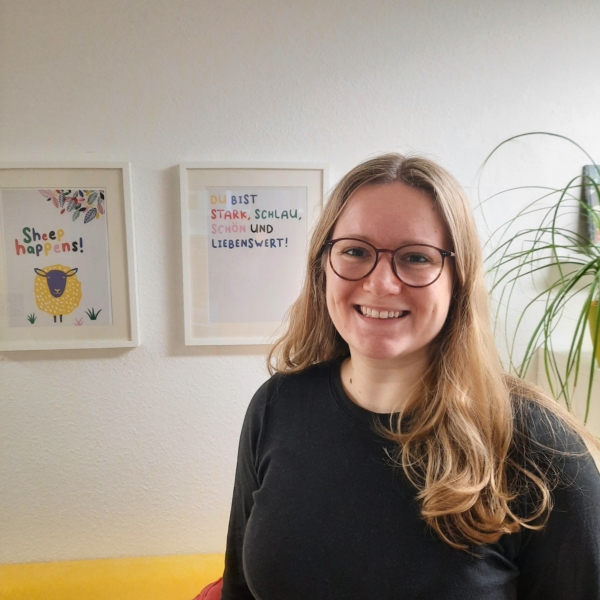 Eine Frau mit langen blonden Haaren und Brille steht lächelnd in einem Wohnzimmer mit gerahmter Kunst an der Wand und einer hohen Pflanze im Hintergrund. Sie trägt einen dunklen Pullover und steht neben einer gelben Couch.