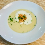Eine weiße Schale mit cremiger Suppe, garniert mit einem Klecks Sahne, einer kleinen Portion Räucherlachs und etwas gehackten Kräutern, auf einem hellen Holztisch.