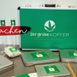 Ein grüner Koffer mit der Aufschrift "Der grüne Koffer" und einem Cannabisblatt, umgeben von Metalldosen, Probengläsern, Reagenzgläsern und Karten, die alle auf einem Tisch vor einem weißen Hintergrund angeordnet sind.