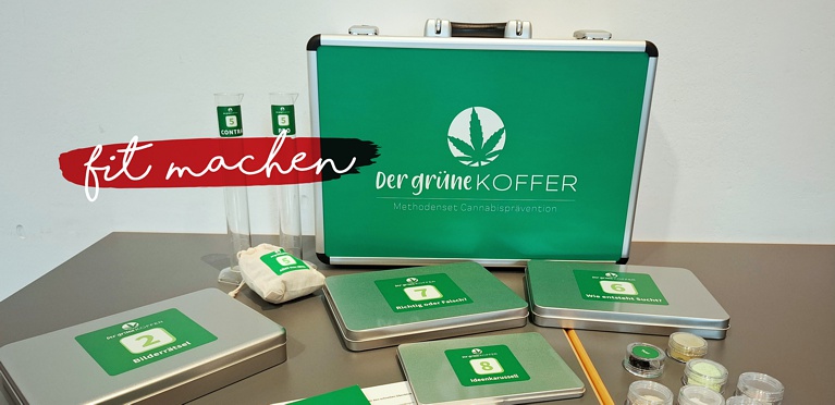 Ein grüner Koffer mit der Aufschrift "Der grüne Koffer" und einem Cannabisblatt, umgeben von Metalldosen, Probengläsern, Reagenzgläsern und Karten, die alle auf einem Tisch vor einem weißen Hintergrund angeordnet sind.