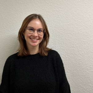 Eine junge Frau mit schulterlangen braunen Haaren, Brille und schwarzem Pullover steht lächelnd vor einer strukturierten, cremefarbenen Wand.
