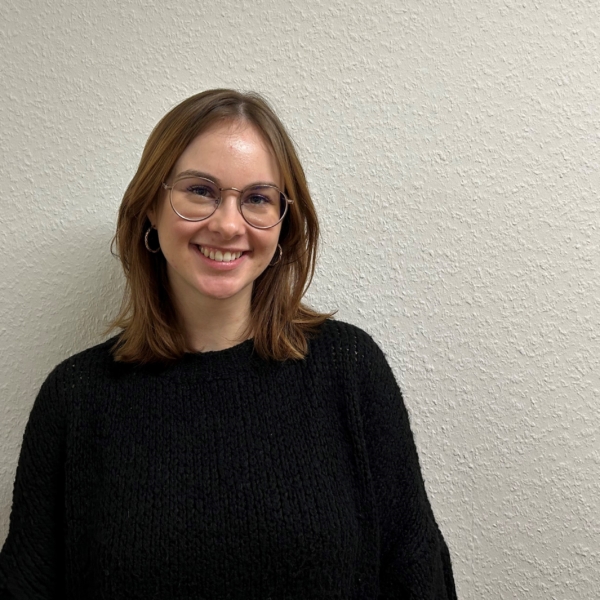 Eine junge Frau mit schulterlangen braunen Haaren, Brille und schwarzem Pullover steht lächelnd vor einer strukturierten, cremefarbenen Wand.