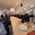 Eine Gruppe von Menschen, meist Männer, steht und sitzt um einen langen Holztresen in einem hell erleuchteten Raum und hört einem Redner aufmerksam zu. Papiere, Servietten und eine Kaffeekanne stehen auf dem Tresen.