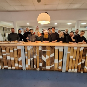 Eine Gruppe von fünfzehn Personen steht lächelnd hinter einer rustikalen Holztheke in einem hell erleuchteten Raum. Auf der Theke stehen Tabletts mit Lebensmitteln, und in der Mitte steht eine Weihnachtsmannfigur, die auf eine festliche Zusammenkunft oder Veranstaltung hindeutet.