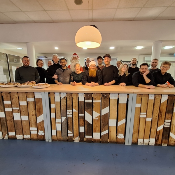 Eine Gruppe von fünfzehn Personen steht lächelnd hinter einer rustikalen Holztheke in einem hell erleuchteten Raum. Auf der Theke stehen Tabletts mit Lebensmitteln, und in der Mitte steht eine Weihnachtsmannfigur, die auf eine festliche Zusammenkunft oder Veranstaltung hindeutet.