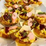 Eine Platte mit getoasteten Brotscheiben, belegt mit geschmolzenem Käse, Mais, in Scheiben geschnittener Wurst, geraspelter Roter Bete und frischem Mikrogrün, angerichtet auf einem weißen Tablett zum Servieren.
