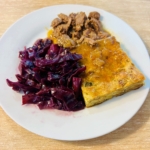 Ein weißer Teller mit geschmortem Rotkohl, in Scheiben geschnittenes gekochtes Fleisch mit Soße und eine rechteckige Scheibe eines herzhaften Auflaufs oder einer Auflaufform auf einem hellen Holztisch.
