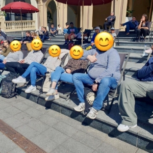 Eine Gruppe von Menschen sitzt auf einer Treppe im Freien und entspannt sich im Sonnenlicht. Die meisten von ihnen tragen Freizeitkleidung und haben Rucksäcke dabei. Andere Menschen sitzen oder gehen im Hintergrund. Alle Gesichter sind mit Smiley-Emojis bedeckt.