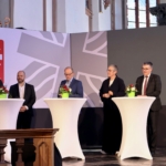 Sechs Personen in förmlicher Kleidung stehen hinter hohen runden Tischen mit Blumenarrangements und sprechen bei einer Veranstaltung. Hinter ihnen sind Logos der Caritas und ein Schild mit der Aufschrift 100 Jahre Caritas Aachen zu sehen. Der Schauplatz scheint eine Kirche oder ein großer Saal zu sein.