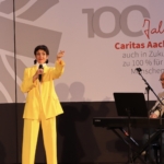 Eine Frau in einem leuchtend gelben Anzug singt auf der Bühne in ein Mikrofon, während ein Mann ein Keyboard spielt. Hinter ihnen hängt ein Banner mit der Aufschrift 100 Jahre Caritas Aachen auch in Zukunft zu 100 % für die Menschen!.
