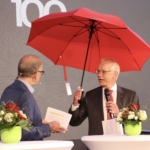 Zwei Männer in Anzügen sprechen auf der Bühne neben runden Tischen mit Blumenarrangements; ein Mann hält einen roten Regenschirm über seinen Kopf. Beide tragen Mikrofone, und im Hintergrund ist eine große "100" zu sehen.