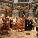 Eine Gruppe von Menschen steht und sitzt auf Holzstühlen in einem großen, kunstvoll verzierten Kirchenraum mit Mosaikböden, gewölbten Säulen und einem großen Kronleuchter. Auf einem Ständer im Vordergrund sind brennende Kerzen aufgestellt.