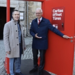 Zwei Männer in formeller Kleidung stehen neben und in einer leuchtend roten Türinstallation im Freien. Auf einem Schild an der Tür steht Caritas öffnet Türen. Im Hintergrund sind ein Steingebäude und Kopfsteinpflaster zu sehen.