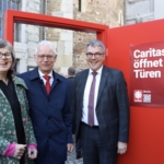 Drei lächelnde Menschen stehen vor einer großen roten Tür mit einem Schild, auf dem Caritas öffnet Türen steht, vor einem historischen Steingebäude.