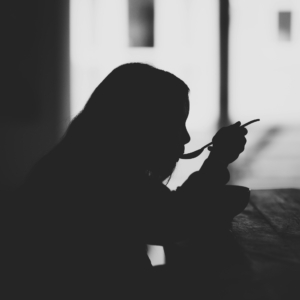 Eine Silhouette einer Person, die an einem Tisch sitzt und einen Löffel an den Mund hält, während sie aus einer Schüssel isst, mit einem hellen Fenster im Hintergrund.