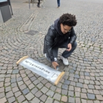 Unser Praktikant Noah sprüht den Link abgezockt.de mit einer Schablone und Kreidefarbe auf den Boden.