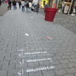 Eine Fußgänger-Einkaufsstraße mit gehenden Menschen. Auf dem Bürgersteig ist die Website "ausgezockt.de" dreimal mit Richtungspfeilen schabloniert. An den Seiten sind Geschäfte und ein großer roter Blumenkübel zu sehen.
