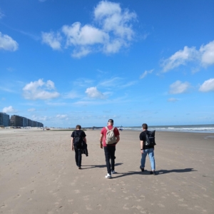 Drei Personen mit Rucksäcken gehen an einem breiten Sandstrand unter einem blauen Himmel mit vereinzelten Wolken entlang. In der Ferne säumen Gebäude die linke Seite, und rechts ist das Meer zu sehen.