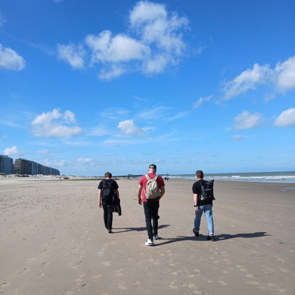 Drei Personen mit Rucksäcken gehen an einem breiten Sandstrand unter einem blauen Himmel mit vereinzelten Wolken entlang. In der Ferne säumen Gebäude die linke Seite, und rechts ist das Meer zu sehen.